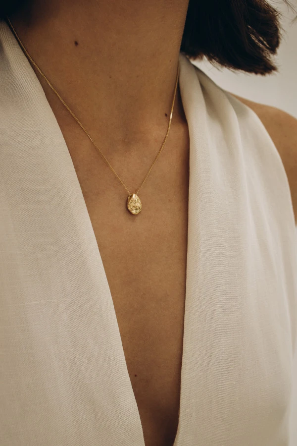 Zbliżenie na zawieszkę SPOT GOLD na szyi modelki – noszona z dekoltem w szpic, złota forma z nieregularną fakturą. Zbliżenie na zawieszkę SPOT GOLD na szyi modelki – noszona z dekoltem w szpic, złota forma z nieregularną fakturą.